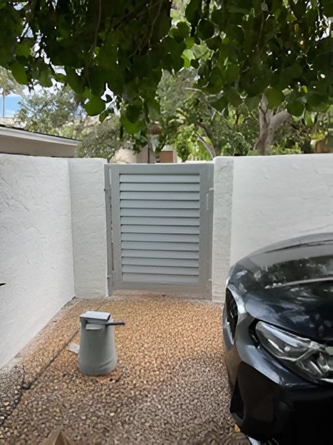 A grey, louvered metal gate set between two white stucco walls on a gravel driveway, with a car visible on the right.
