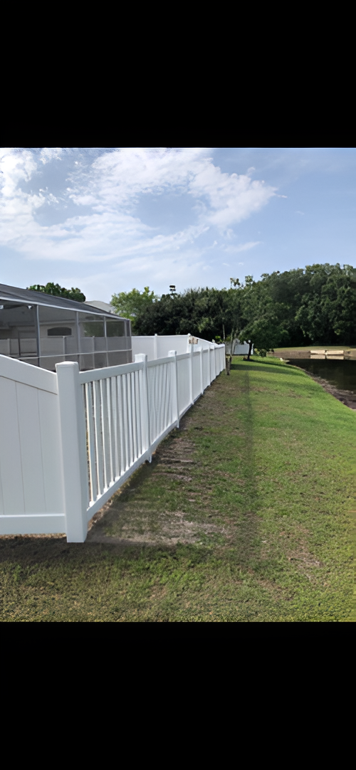 A white vinyl fence runs along the edge of a grassy lawn, with a screened-in pool enclosure and trees in the background.