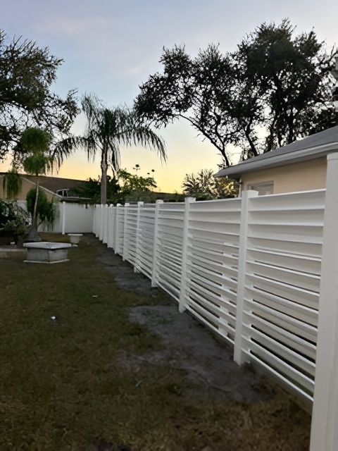 A horizontal white slatted vinyl fence runs through a backyard with trees and a house in the background at sunset.