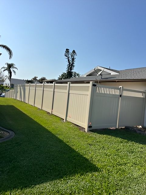 A long, tan-colored vinyl privacy fence stretches across a green residential lawn under a clear blue sky.