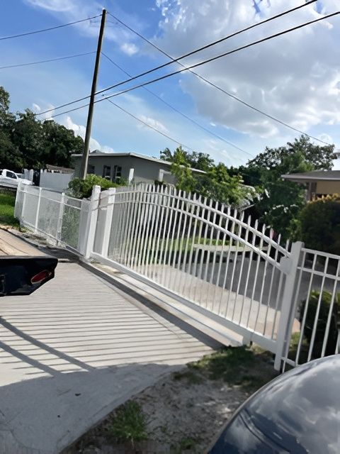 A white metal driveway gate stands closed across a driveway in front of a residential house under a blue, cloudy sky.