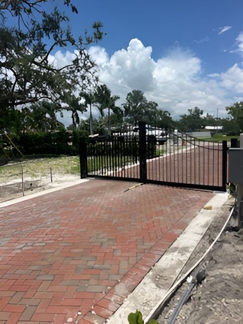 A black metal gate stands across a brick driveway under a sunny sky with palm trees in the background.