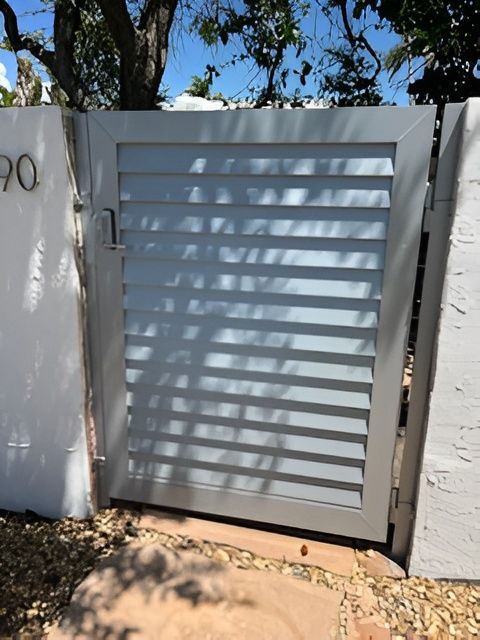 A light gray metal gate with horizontal slats installed between two white stucco pillars in a gravel yard.