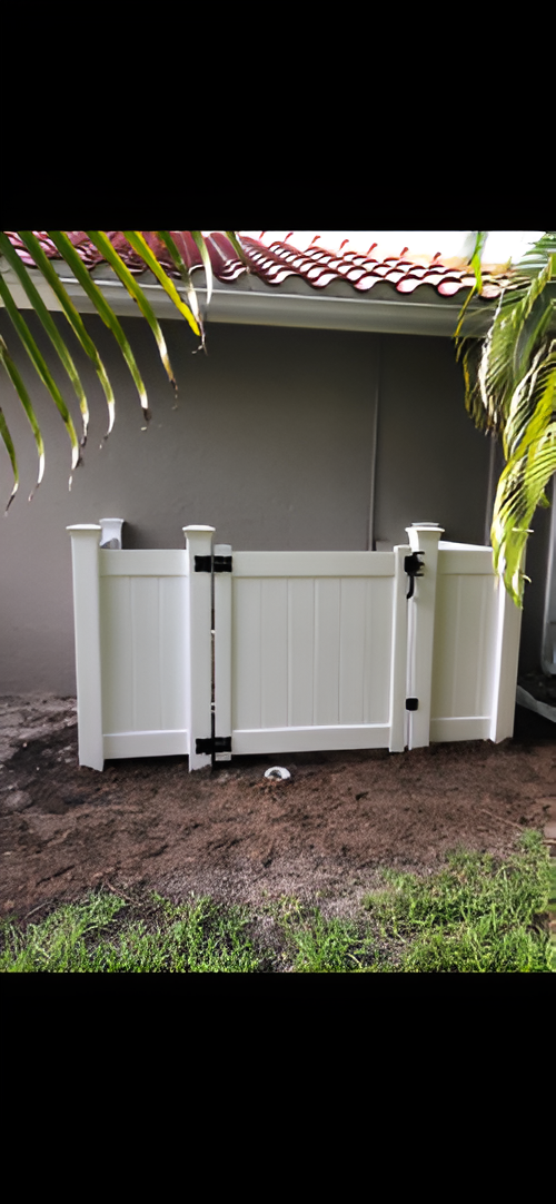 A white vinyl privacy fence panel with a gate, installed against a gray exterior wall and mulch ground.