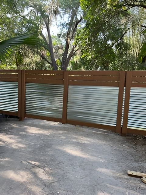 A modern backyard fence featuring horizontal wooden framing with corrugated metal panel inserts, set in a wooded area.