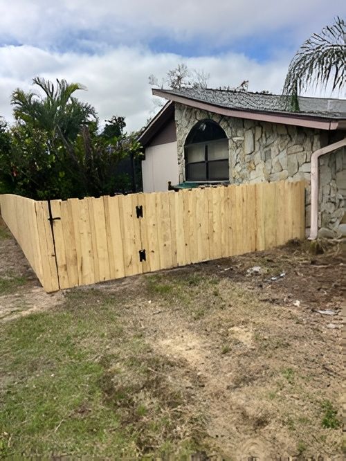 A newly installed wooden privacy fence runs alongside a house with stone-accented siding and a green roof.