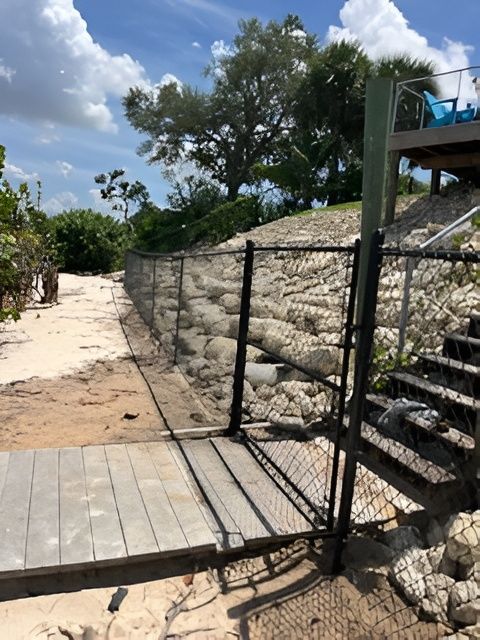 A wooden boardwalk bridge leads to a black chain-link fence and stone embankment next to a sandy beach under a blue sky.