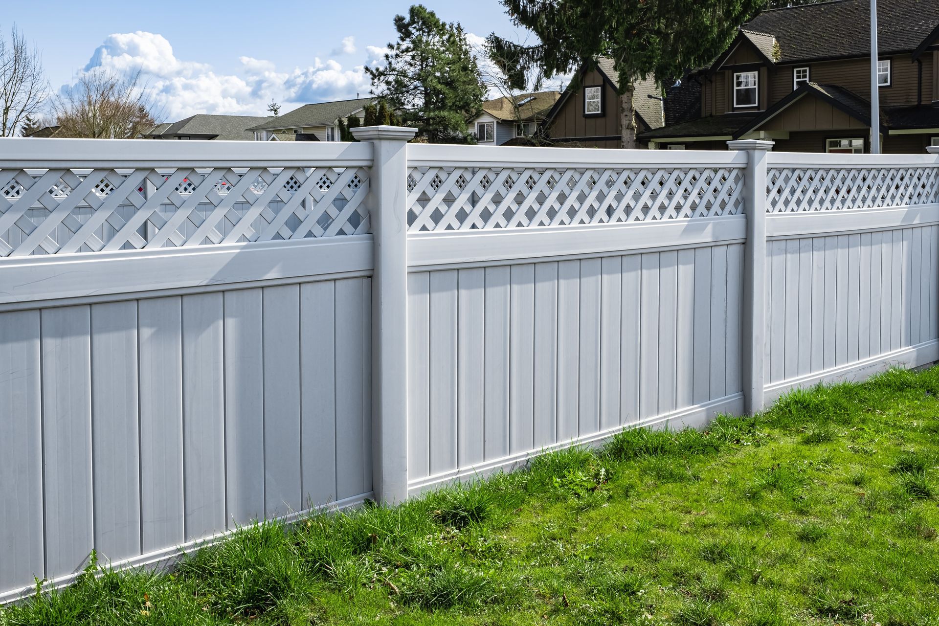 A white vinyl fence with a lattice top border, positioned outdoors along a grass lawn against a suburban backdrop.