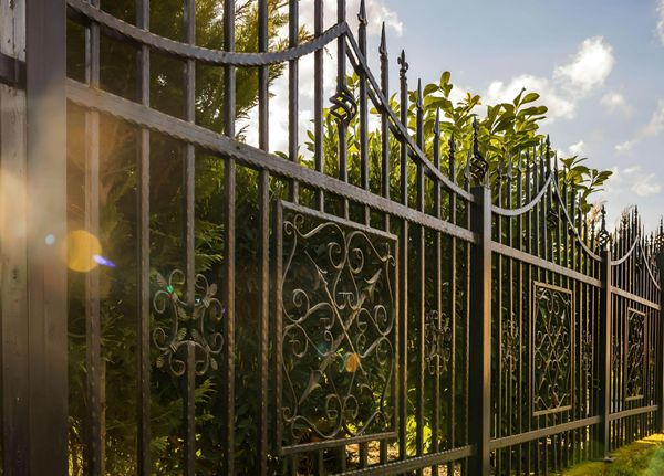 Black wrought-iron decorative fence with scrollwork panels and pointed finials, set against lush green foliage.