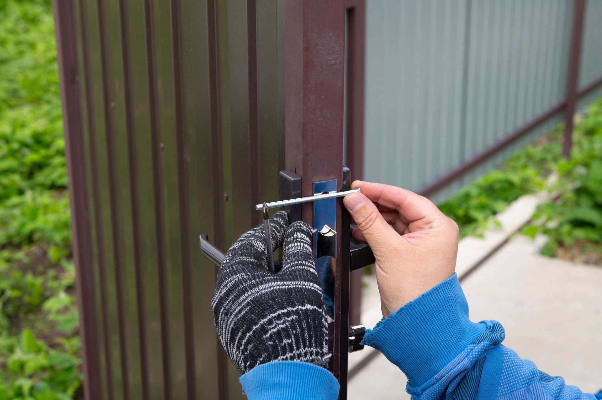 Hands in work gloves install a metal slide bolt lock on a metal fence gate.