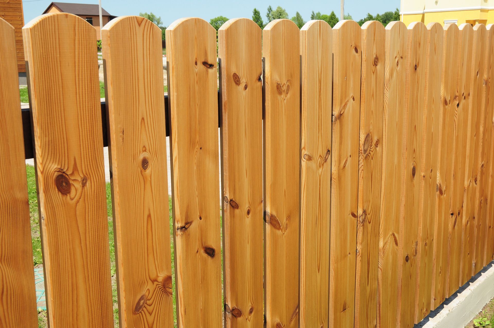 A close-up view of a new, light-brown wooden picket fence with a rounded top design, set against a blurred background.