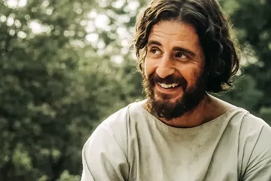 A man with a beard and long hair is smiling. Jesus. Jonathan Roumie.