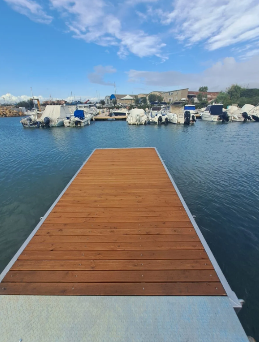Molo in legno che si estende nelle calme acque blu, con barche in un porto sotto un cielo parzialmente nuvoloso.