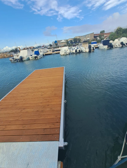 Il molo in legno si estende in acque calme e azzurre, con barche ormeggiate sullo sfondo sotto un cielo azzurro.