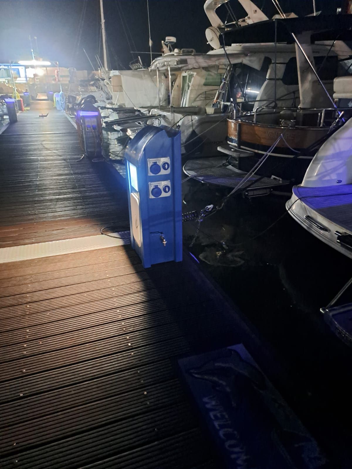Una scatola blu della corrente elettrica su un molo di legno di notte, con barche attraccate nelle vicinanze.