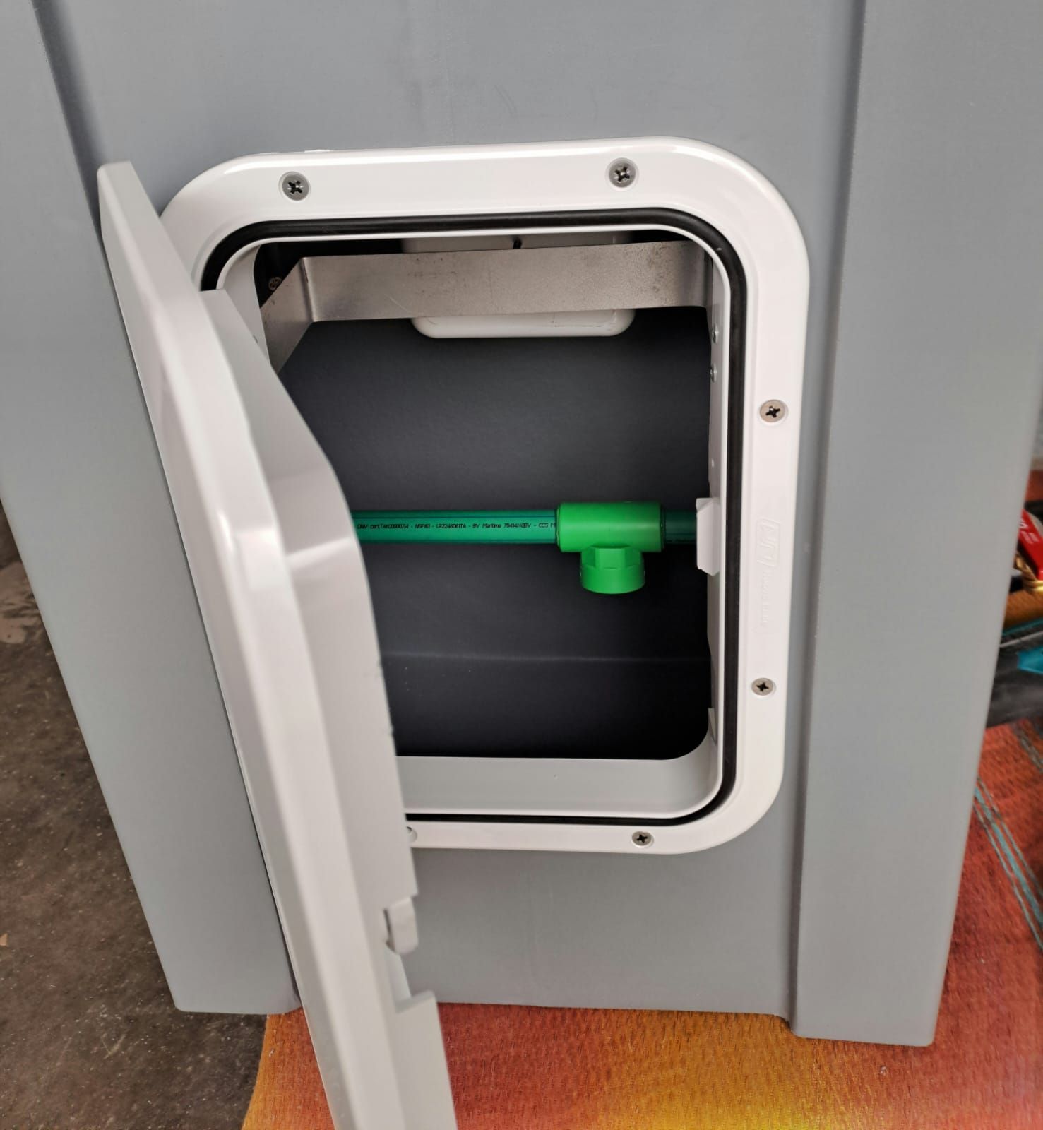 Il pannello di accesso aperto su un contenitore grigio rivela un tubo verde con un raccordo all'interno del vano.