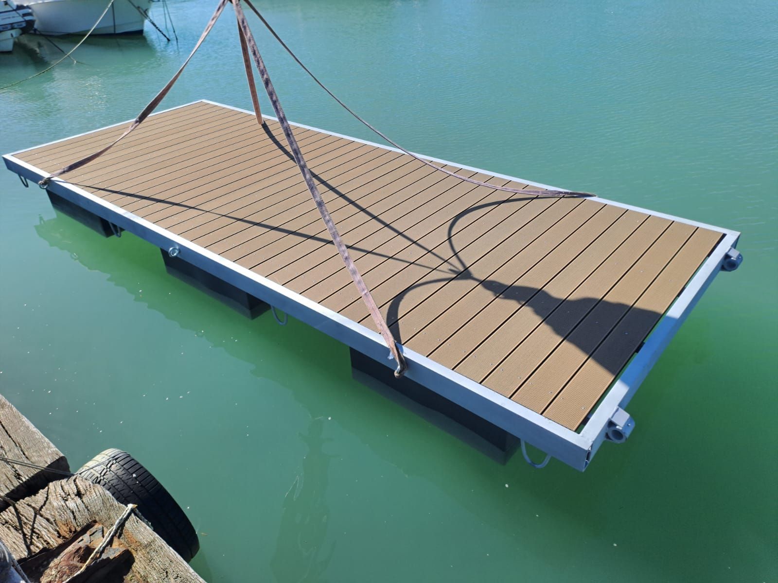 Pontile galleggiante con pavimentazione marrone e struttura grigia, sospeso sull'acqua tramite cinghie.