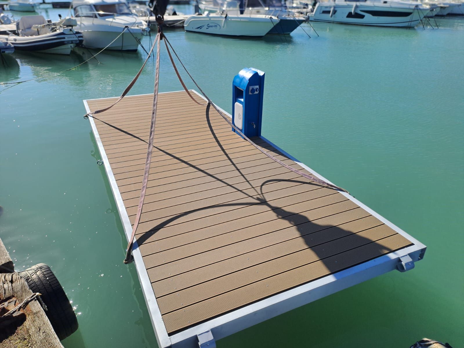 Pontile galleggiante calato in un porto turistico con acqua blu e barche. Pavimentazione marrone, chiosco blu.
