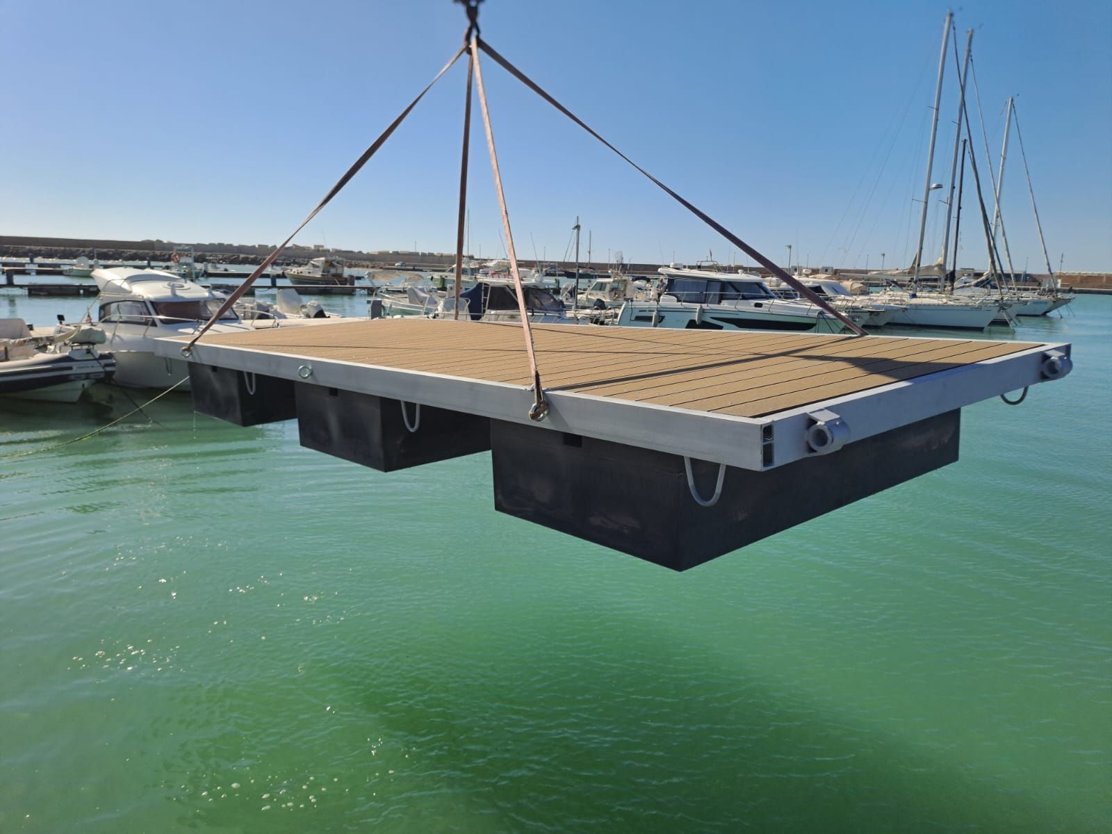 Un molo galleggiante rettangolare viene sollevato tramite cinghie sulle acque turchesi nei pressi di un porto turistico con barche.