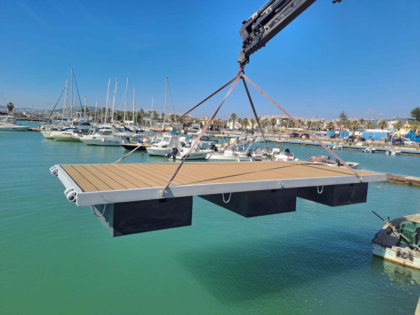 Una gru solleva un grande molo galleggiante con il ponte marrone sopra un porto turistico con barche.