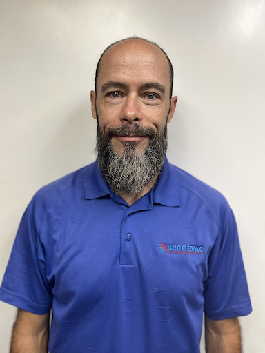 Man with a graying beard, wearing a blue polo shirt, standing in front of a white wall.