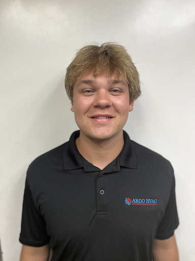 A young person with light brown hair smiles, wearing a black polo shirt with a logo, standing in front of a white wall.