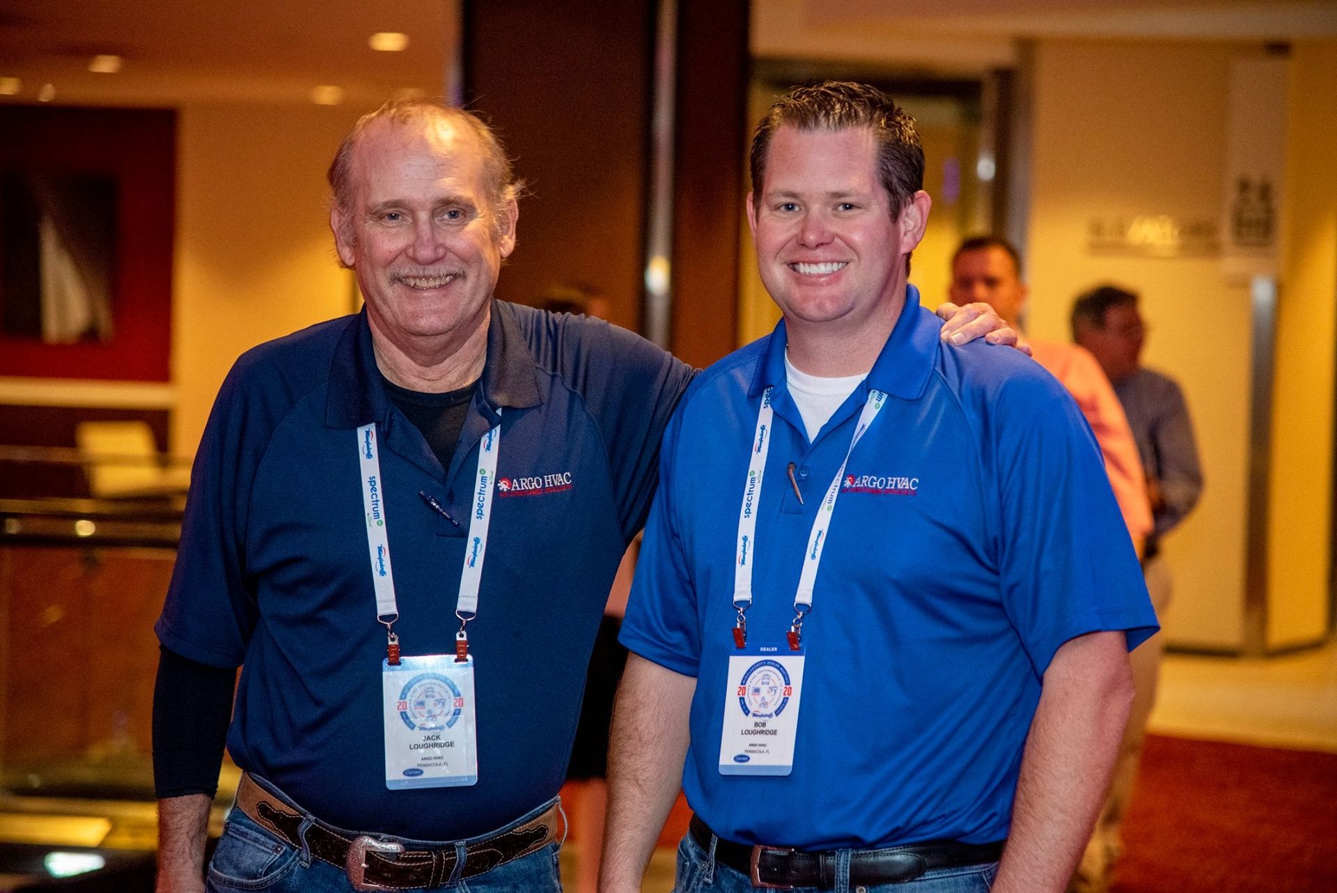 Two men in blue shirts with lanyards smile, standing indoors, presumably at a conference or event.