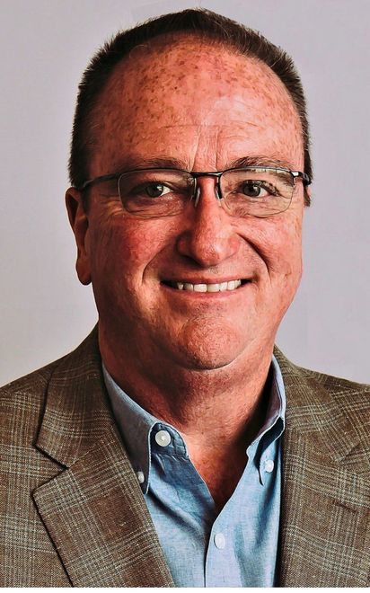 Man with glasses wearing a blue shirt and brown jacket smiles at the camera.
