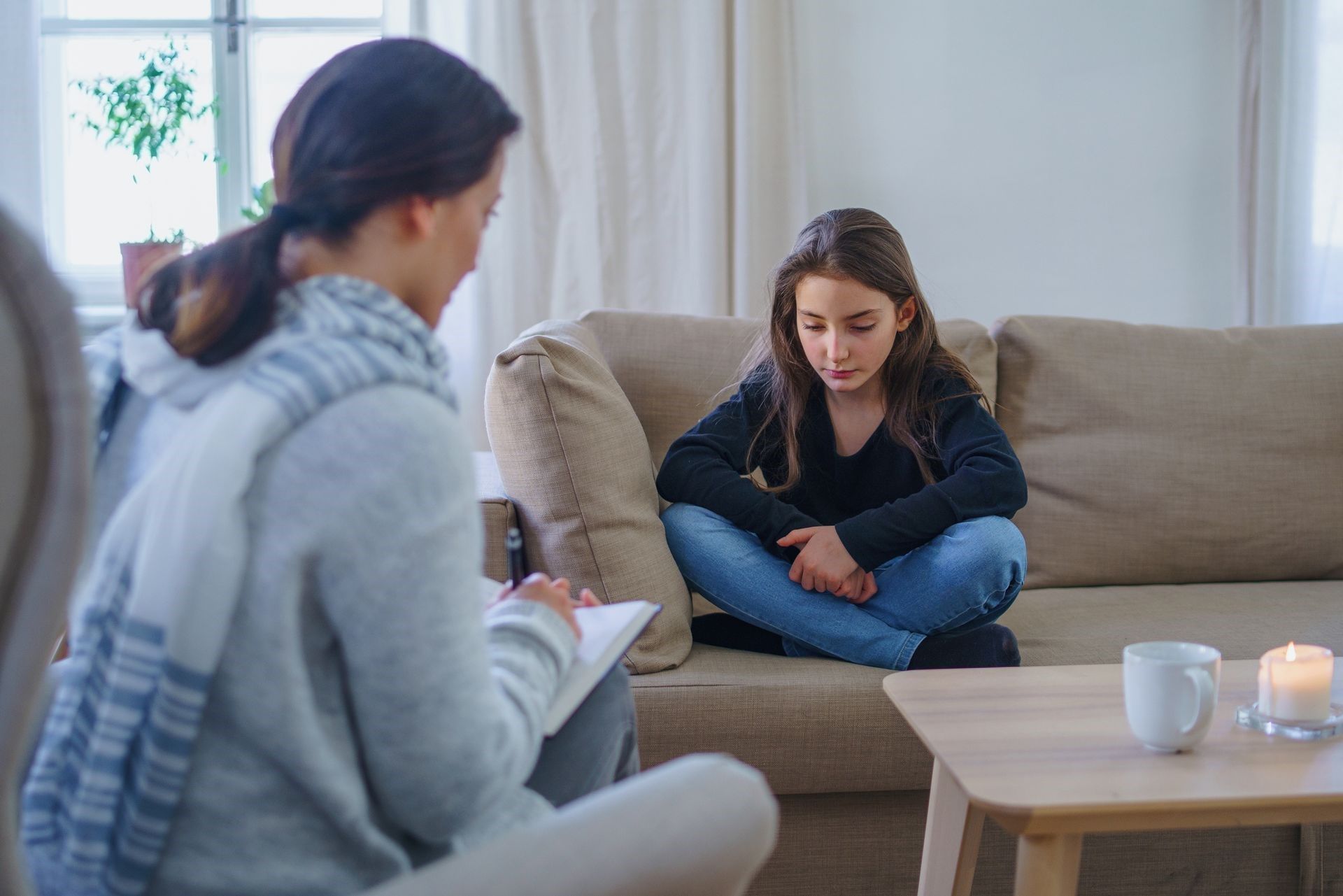 Two people sit on a couch in a bright living room, one writing while the other looks down thoughtfully.