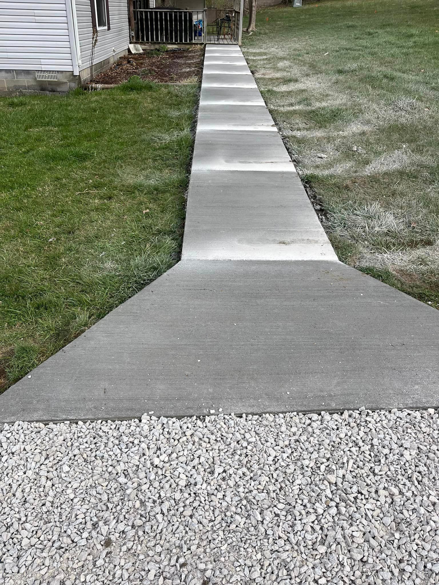 A concrete walkway leading to a house next to a grassy field.
