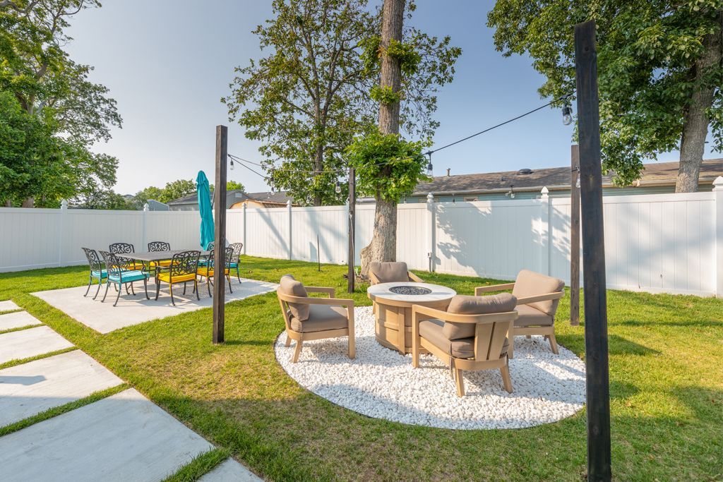 Backyard patio with seating area on white pebbles, dining area with table, grass lawn, and white fence.