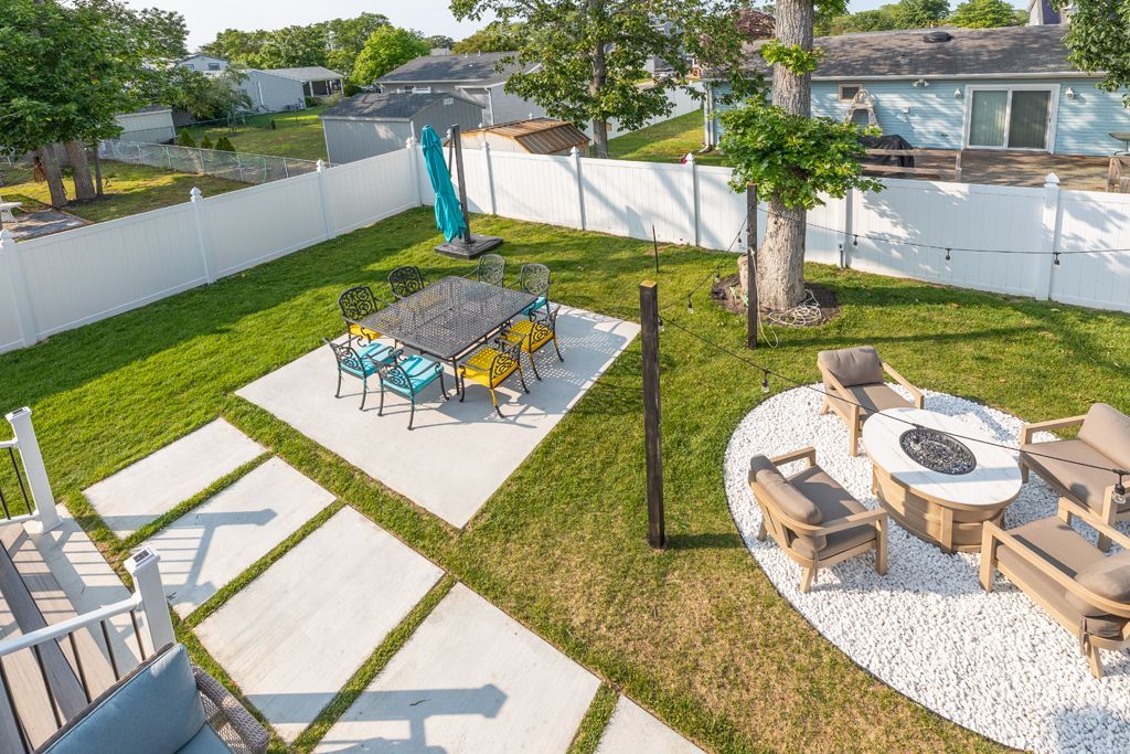 Backyard with patio, lawn, white fence, dining area, and fire pit on decorative stones.