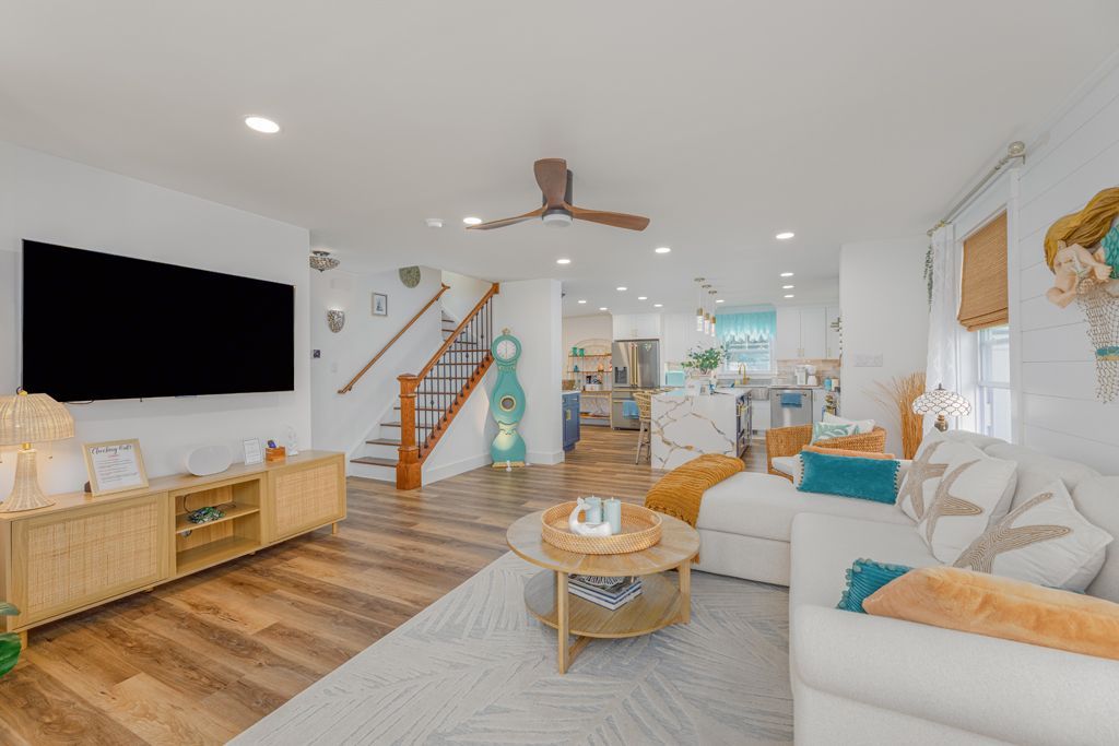 Spacious living room with white walls, wooden floor, and a sectional sofa. Includes a mounted TV, stairs, and kitchen view.