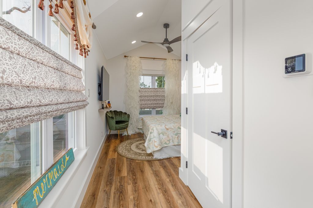 Narrow bedroom with wood floor, white walls, and a view of a bed and a chair.