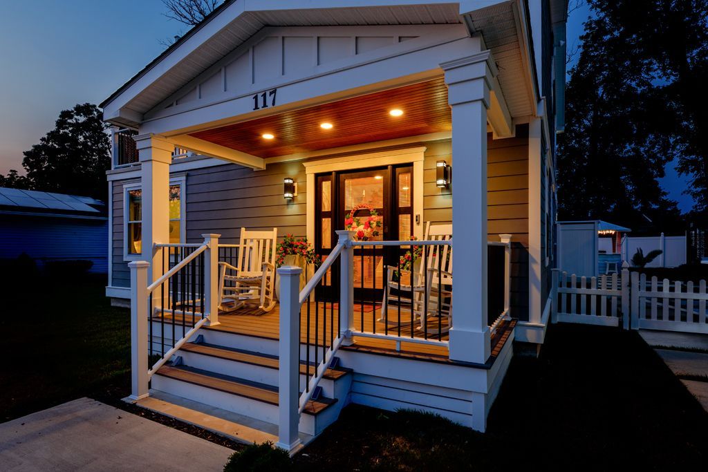 Front porch of a small house at dusk with rocking chairs, lights, and address
