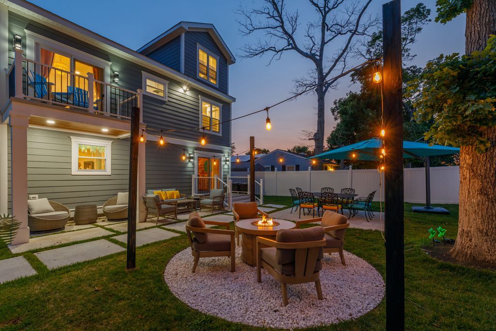 Backyard with seating around fire pit, string lights, and a two-story house at dusk.