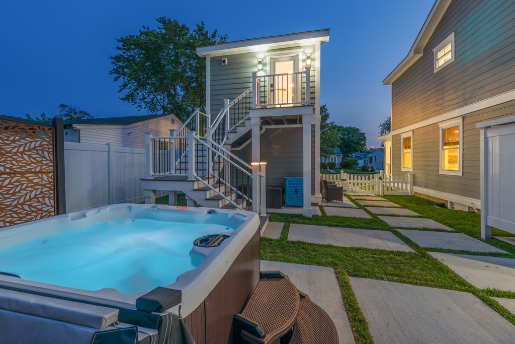 Backyard at dusk with lit hot tub, spiral staircase to elevated room, and paved walkway.