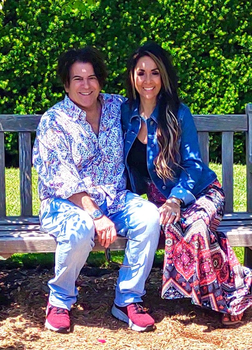 Couple seated on a wooden bench, smiling. Man in patterned shirt and jeans. Woman in denim jacket and patterned skirt. Outdoors, sunny.