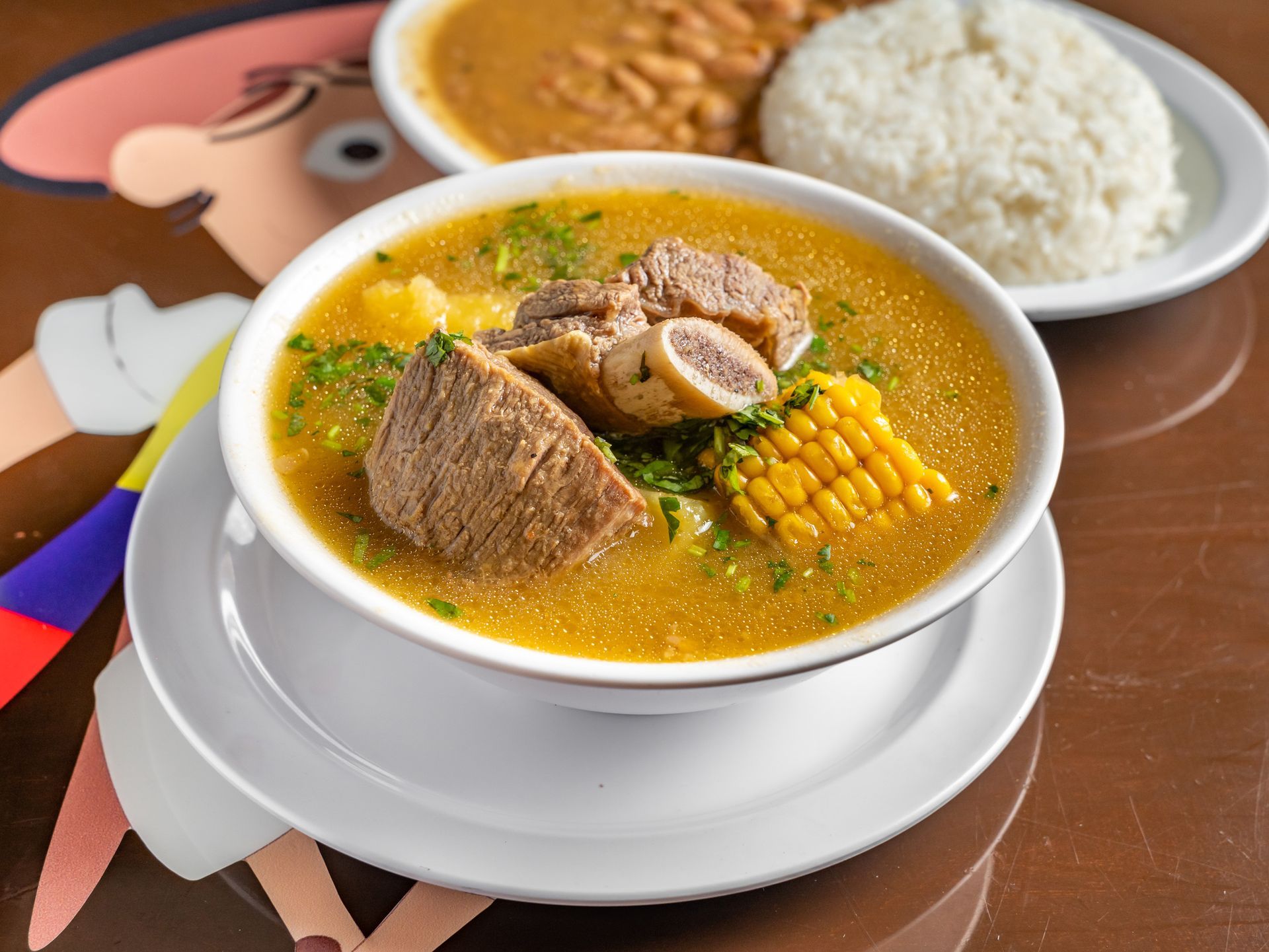 A bowl of soup with meat and corn on the cob next to a plate of rice.
