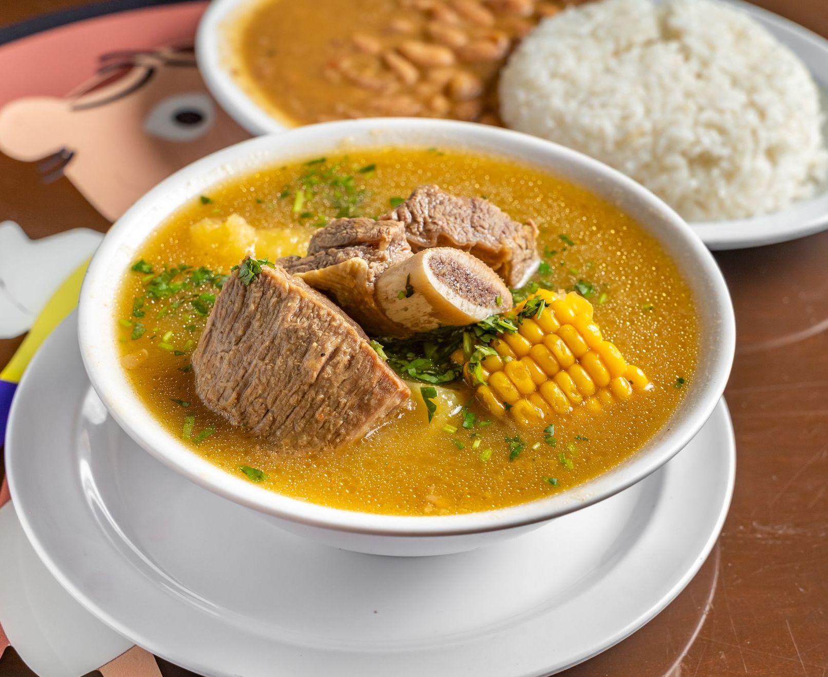 A bowl of soup with meat and corn on the cob next to a plate of rice