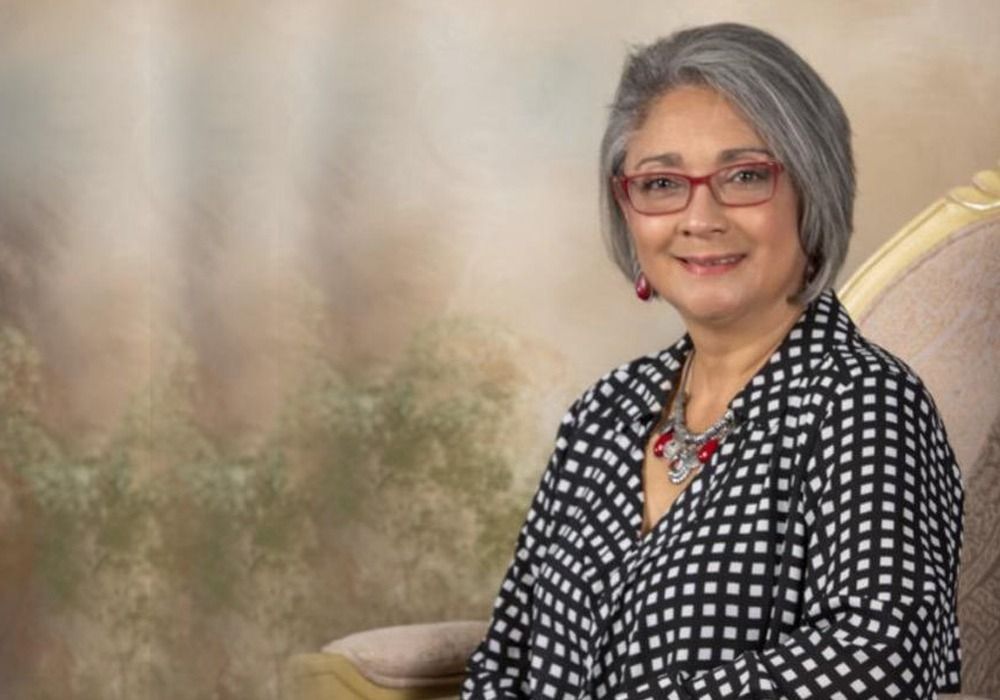 Smiling woman with gray hair and glasses in a checkered shirt, seated in a chair.