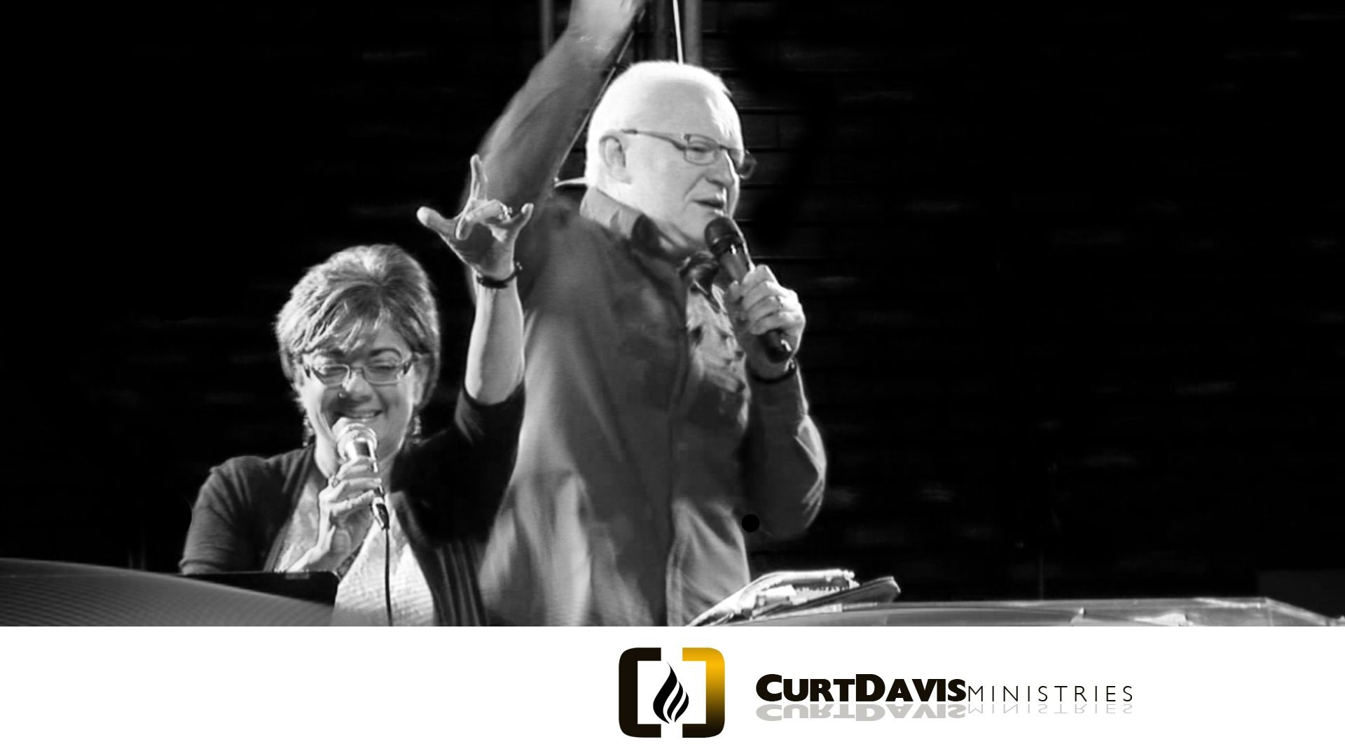 Two people on stage, a man with glasses raising arm, and a woman singing with a microphone. Black and white.