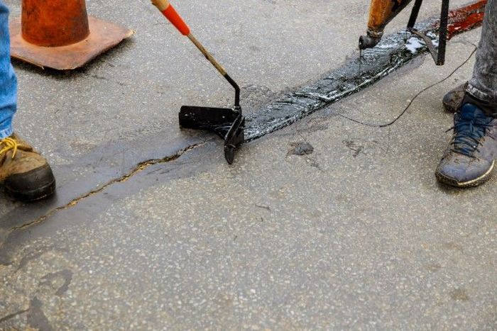 Person filling a crack in pavement with black sealant using a roller tool, next to a traffic cone.