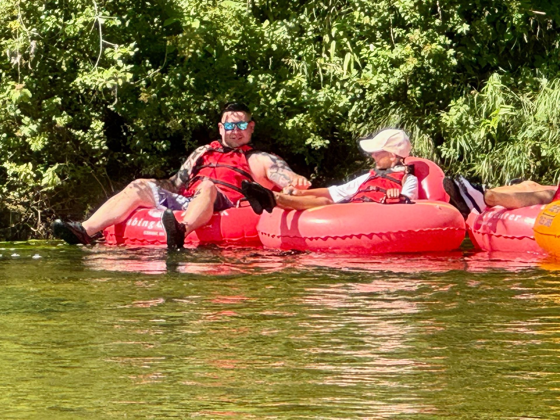 Two men are floating on red inner tubes in a river.
