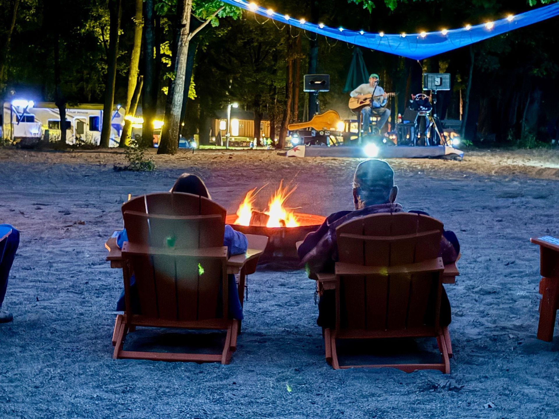 Two people are sitting in chairs in front of a fire pit watching a concert.