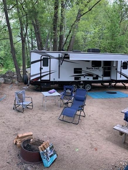A rv is parked in a campground with chairs and a fire pit.