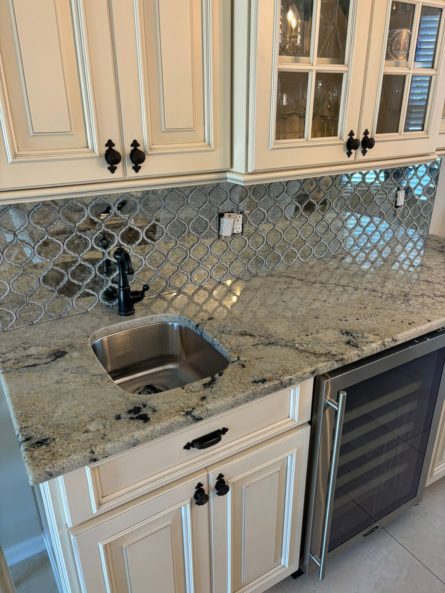 A small kitchen bar area with granite countertop, stainless steel sink, wine fridge, and decorative backsplash.