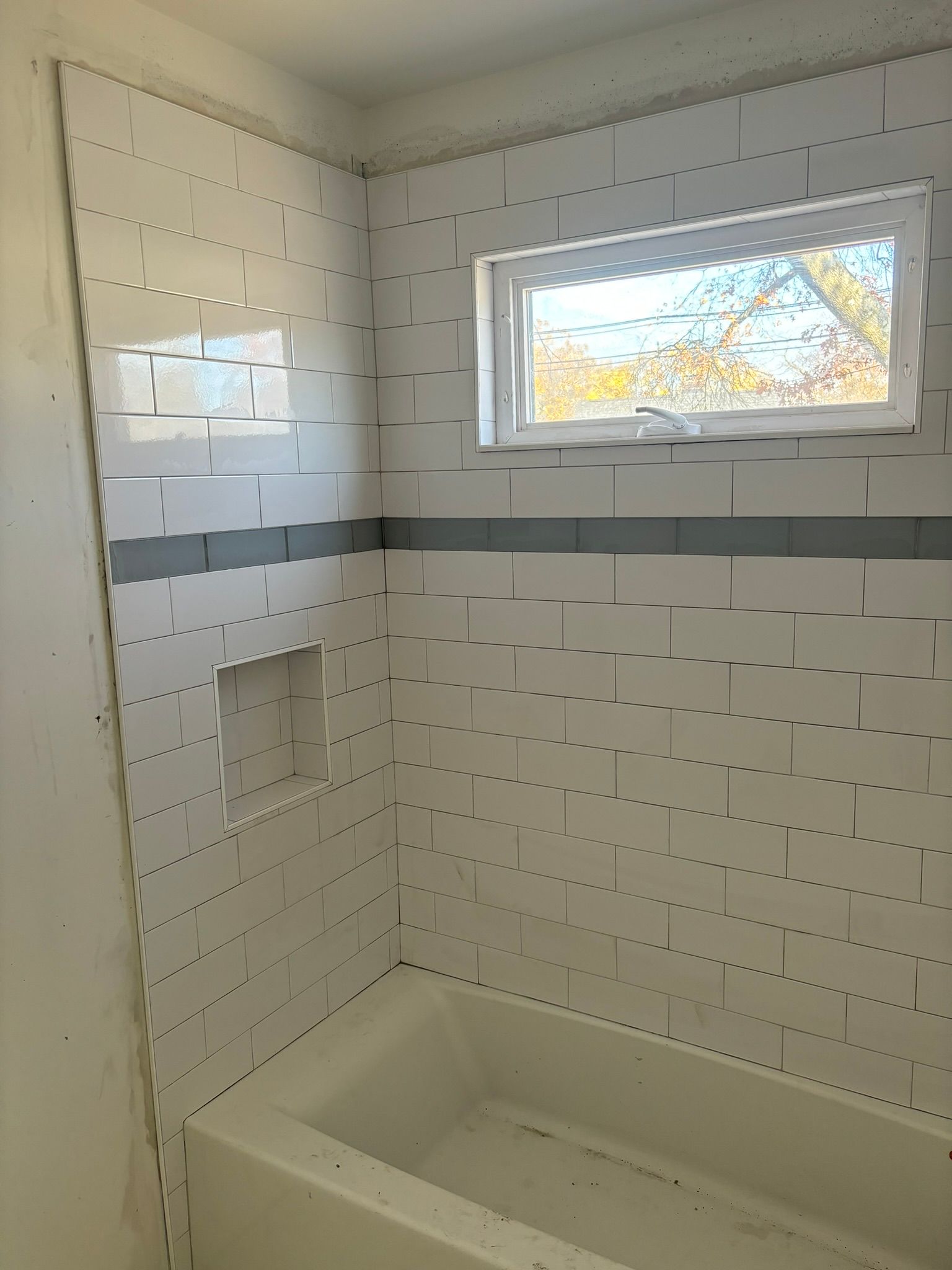 Bathroom with white tiled walls, a window, and a bathtub. Gray stripe accent and a recessed shelf.