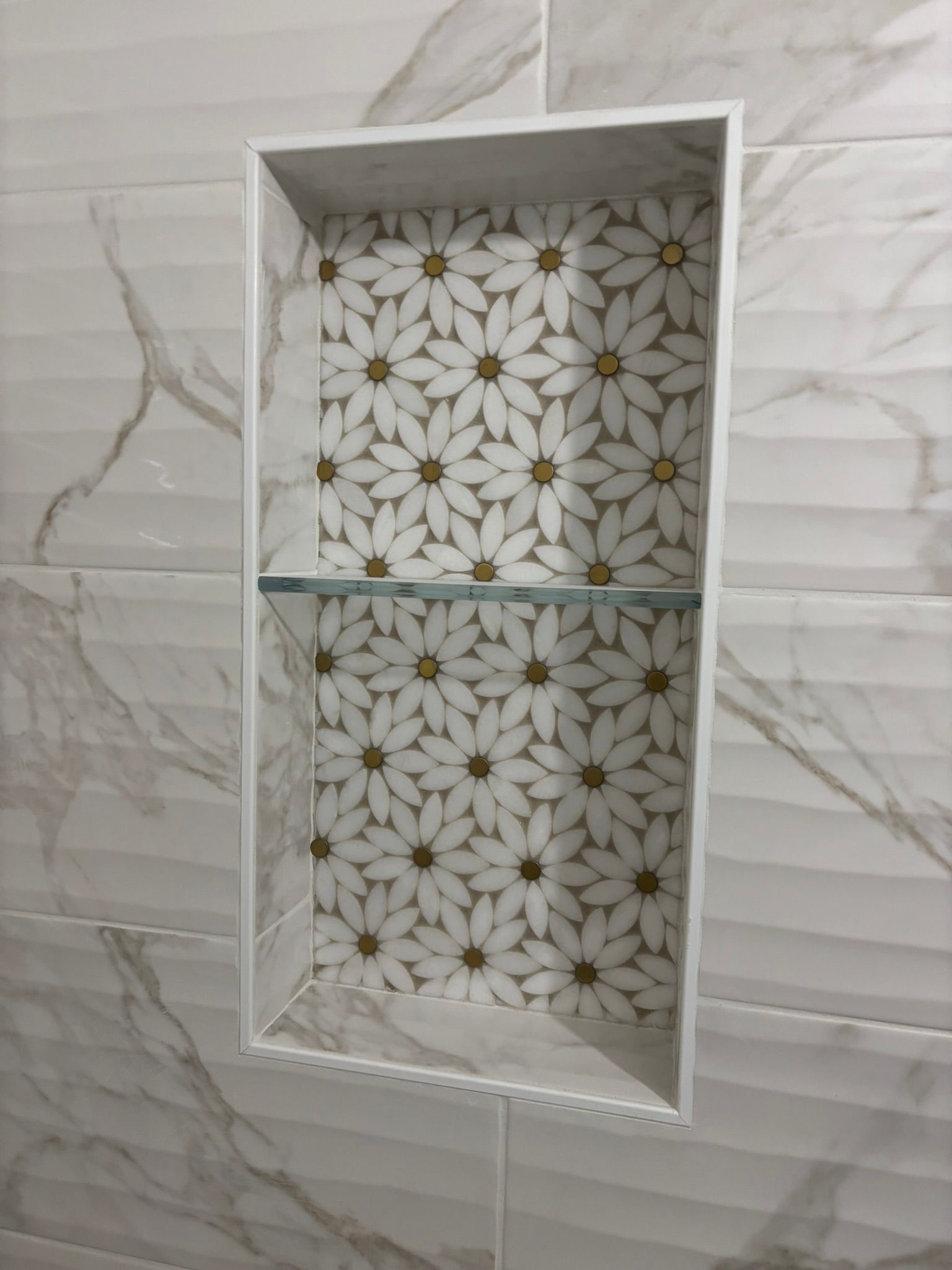 Rectangular tiled shower niche with daisy patterned tile backing, glass shelf. White frame on a wall of marbled tiles.