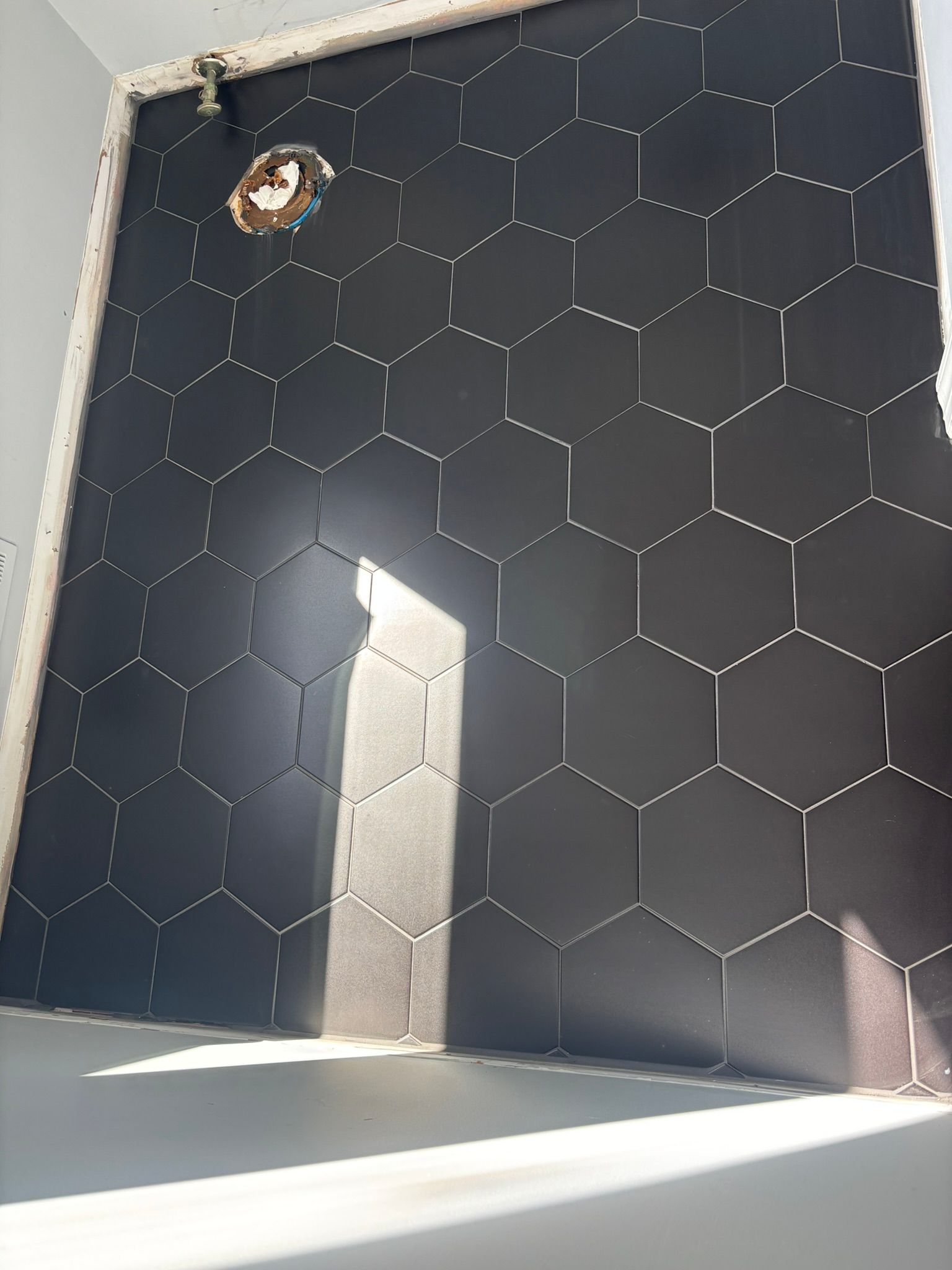 Black honeycomb tiles cover a wall; a pipe is visible in the top center. Light shines onto a gray floor.